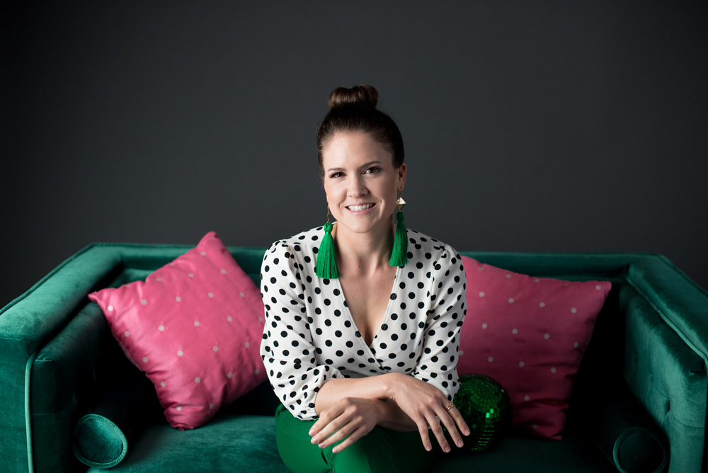 Michelle Grasek, acupuncturist and marketing expert, sitting on a green couch with pink cushions, ready to show you how to get a steady stream of new patients in your clinic.