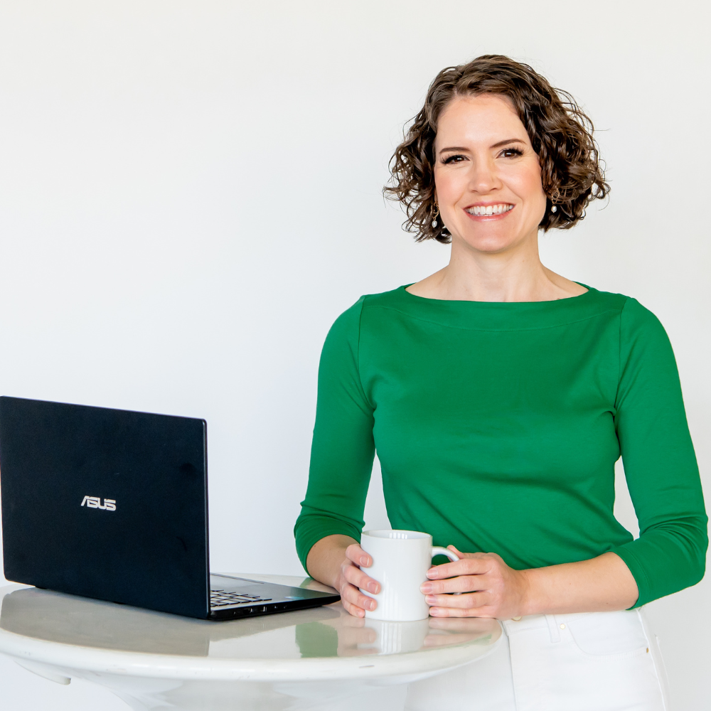 Portrait of Michelle Grasek, host of the Acupuncture Marketing School podcast, wearing a green shirt standing at a desk with a laptop.