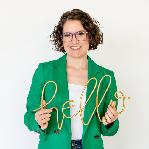 Acupuncturist, marketing strategist, and host of the Acupuncture Marketing School Podcast, Michelle Grasek, wearing a green blazer and purple glasses holding a sign that says Hello.